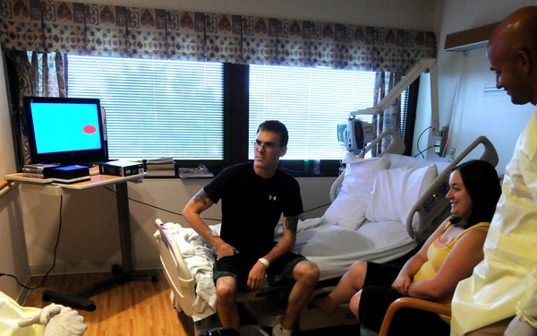 A hospital room scene showing four individuals engaged in conversation. Two people are wearing yellow protective gowns and gloves, suggesting infection control protocols are in place. The setting appears to be a patient room with medical equipment, a hospital bed, and personal items visible.