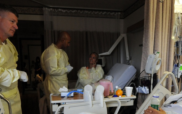 A hospital room scene showing a young male patient lying in a bed while being visited by medical personnel wearing yellow protective gowns and white gloves. At least three staff members are present in the room, with one engaging directly with the patient. Medical equipment including an IV stand, monitoring devices, and a bedside tray with food items are visible.