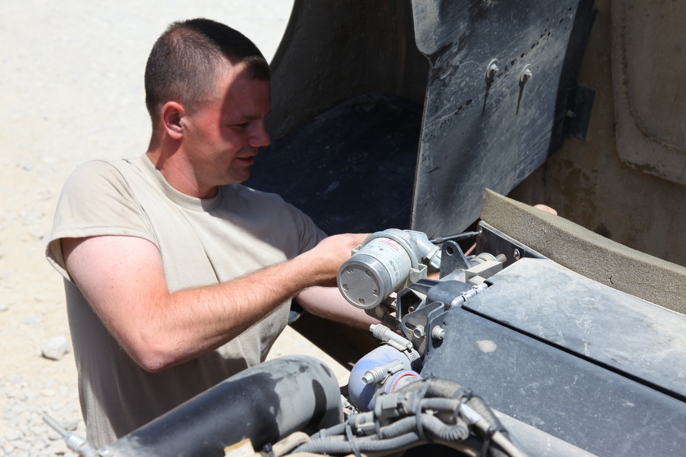 A male individual wearing a tan/beige military-style t-shirt is performing mechanical maintenance on a military vehicle. He is leaning into an open engine compartment or access panel, handling what appears to be a cylindrical component, likely a filter or motor assembly. The setting appears to be an arid, outdoor environment consistent with a forward operating base or desert location.