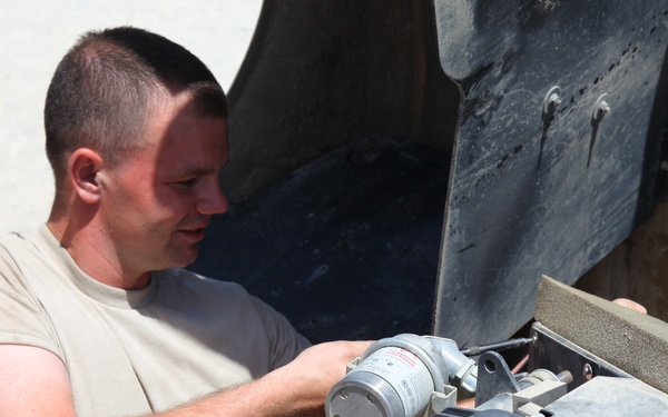 A male individual wearing a tan/beige military-style t-shirt is performing mechanical maintenance on a military vehicle. He is leaning into an open engine compartment or access panel, handling what appears to be a cylindrical component, likely a filter or motor assembly. The setting appears to be an arid, outdoor environment consistent with a forward operating base or desert location.