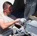 A male individual wearing a tan/beige military-style t-shirt is performing mechanical maintenance on a military vehicle. He is leaning into an open engine compartment or access panel, handling what appears to be a cylindrical component, likely a filter or motor assembly. The setting appears to be an arid, outdoor environment consistent with a forward operating base or desert location.