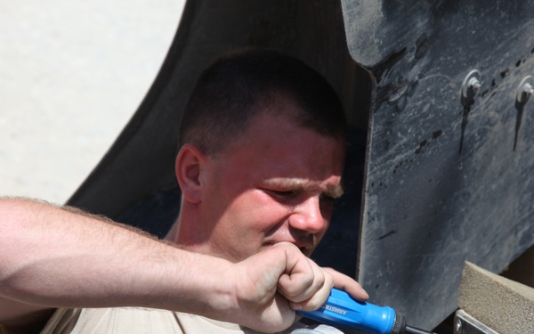 A young male in a tan/beige military-style shirt is performing maintenance work on a military vehicle, using a blue screwdriver on a cylindrical component. He is crouched or leaning under the vehicle's wheel well area in what appears to be a desert or arid environment. The work appears to involve a mechanical or electrical component mounted on the vehicle's undercarriage or lower chassis.