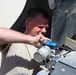 A young male in a tan/beige military-style shirt is performing maintenance work on a military vehicle, using a blue screwdriver on a cylindrical component. He is crouched or leaning under the vehicle's wheel well area in what appears to be a desert or arid environment. The work appears to involve a mechanical or electrical component mounted on the vehicle's undercarriage or lower chassis.