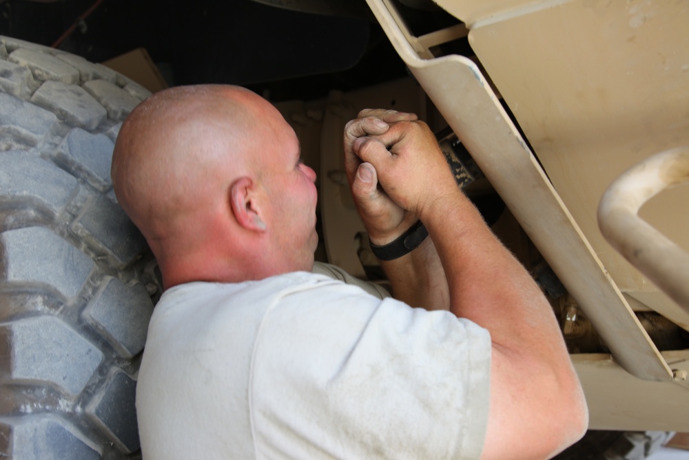 A bald man in a white t-shirt is lying on his back performing mechanical work underneath a large military-style vehicle, likely a HMMWV (Humvee) based on the tan/desert-colored body panels and large off-road tire. His hands are visibly dirty and he is working on components in a tight space between the wheel well and undercarriage. The setting appears to be an outdoor or semi-covered maintenance area.