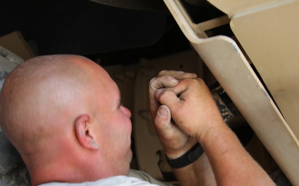 A bald man in a white t-shirt is lying on his back performing mechanical work underneath a large military-style vehicle, likely a HMMWV (Humvee) based on the tan/desert-colored body panels and large off-road tire. His hands are visibly dirty and he is working on components in a tight space between the wheel well and undercarriage. The setting appears to be an outdoor or semi-covered maintenance area.