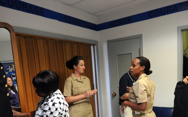 The image shows a group of individuals in what appears to be a military uniform fitting or inspection room. Several personnel in tan/khaki Navy uniforms are being assisted by civilian helpers with uniform adjustments and fittings. The room has motivational words such as 'COURAGE,' 'HONOR,' and 'COMMITMENT' displayed as a border near the ceiling.
