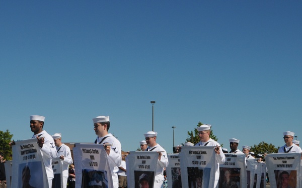 U.S. Navy sailors in dress white uniforms are marching in a parade while carrying large memorial banners displaying photographs and information about fallen service members. The banners appear to honor military personnel who died in service, with names, ranks, branches, dates, and hometowns visible on each panel. The event appears to be a Memorial Day or Veterans Day parade held on a clear, sunny day.