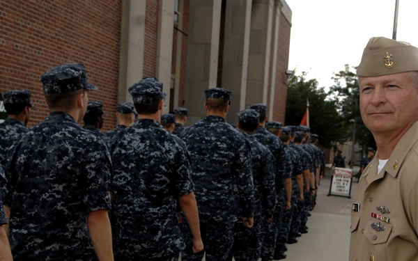 A senior U.S. Navy officer in tan Service Dress Khaki uniform stands at attention while observing a column of enlisted sailors marching in Navy Working Uniform (NWU Type I digital blue camouflage). The scene appears to take place on a military installation or naval base, with brick buildings and flagpoles visible in the background. The officer appears to be overseeing or inspecting the formation as it passes.