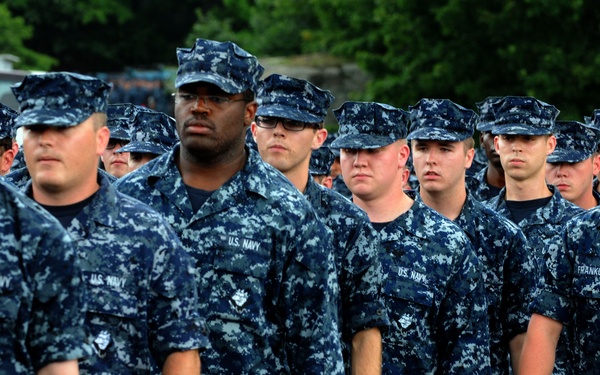 A large formation of U.S. Navy personnel standing at attention in Navy Working Uniform (NWU) Type I, characterized by the blue digital camouflage pattern. The sailors appear to be participating in a formal military formation or ceremony outdoors. Multiple rows of personnel are visible, with trees and a brick building in the background.