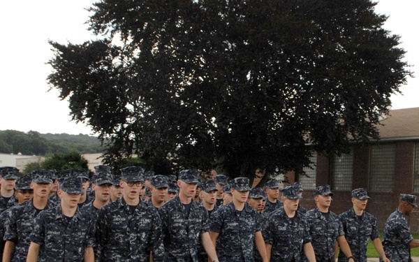 A large formation of U.S. Navy personnel in Navy Working Uniform (NWU) Type I digital blue camouflage are marching in formation along a paved road. A senior officer or petty officer in khaki uniform stands to the right, appearing to direct or observe the formation. The setting appears to be a military installation with brick buildings and wooded hills in the background.