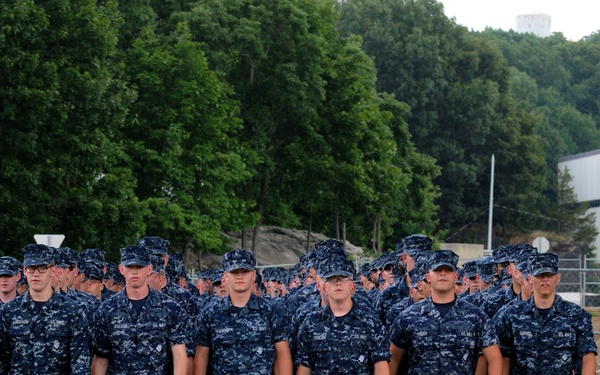 A large formation of U.S. Navy personnel marching in formation on a paved road or parade ground. All personnel are wearing the Navy Working Uniform (NWU) Type I, characterized by its blue digital camouflage pattern. A single sailor appears to be leading or flanking the formation on the right side.
