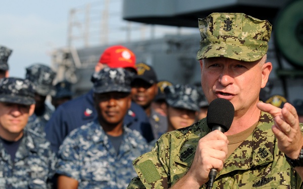 A U.S. Navy officer in olive/green digital camouflage (AOR2 pattern) is addressing a group of sailors aboard what appears to be a naval vessel. He is speaking into a handheld microphone while gesturing with his right hand, suggesting an official address or briefing. The assembled sailors behind him are wearing the standard Navy Working Uniform (NWU Type I) blue digital camouflage pattern.