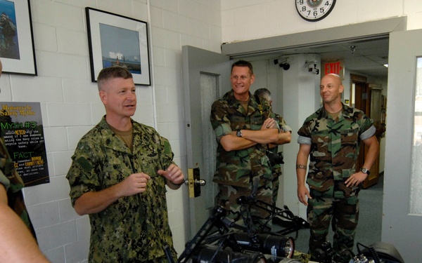 Military personnel in a briefing or demonstration room are gathered around a table displaying what appear to be small ground robots or remotely operated vehicles. Four individuals are visible, wearing U.S. Navy working uniform (NWU) and U.S. military woodland camouflage (BDU) patterns. The setting appears to be an indoor military facility with framed photos, safety posters, and a wall clock visible.
