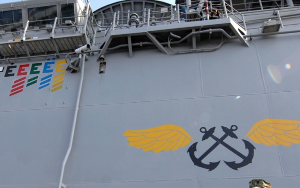 A senior military officer in camouflage uniform addresses a group of Navy personnel gathered on or near a naval vessel. The ship's hull displays a large Naval Aviation insignia (crossed anchors with wings) and multiple 'E' excellence award pennants in various colors. The setting appears to be a pier-side ceremony or briefing.