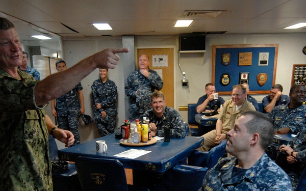 A senior military officer in camouflage uniform is addressing a group of Navy personnel seated at tables in what appears to be a shipboard mess or briefing room. The officer is pointing and appears to be speaking animatedly, with the audience visibly engaged and smiling. The setting includes display boards with military insignia, plaques, and awards mounted on the walls.