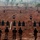A military or law enforcement shooting range exercise is taking place on a red-dirt open field. Multiple personnel in camouflage uniforms stand in a line facing rows of silhouette targets at varying distances. The setting appears to be in a semi-arid or savanna environment based on the sparse, dry vegetation in the background.
