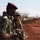 Two soldiers in different uniforms are engaged in what appears to be a military training or coordination exercise in an arid, red-soil terrain. One soldier wears green camouflage with a maroon beret, while the other wears desert-tan combat gear with a helmet and goggles. The soldier in tan gear is pointing in a specific direction, suggesting tactical communication or instruction.