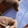 A person wearing tan/desert-colored tactical gloves is holding a spiral notepad with hand-drawn sketches and handwritten text. The notepad contains what appears to be a diagram or schematic drawing along with text that reads 'BZD' and 'SD-100' or similar notation. The setting appears to be outdoors on red/clay soil terrain.
