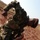 A soldier in camouflage uniform and a dark beret is crouched low to the ground on red laterite soil, appearing to examine or interact with something on the ground surface. The setting is an open, arid terrain with sparse dry grass and red earth, consistent with sub-Saharan African landscape. The image is taken from a low angle with some motion blur on the upper body.