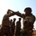 Military personnel in camouflage uniforms and brown berets are engaged in a hand-to-hand combat training exercise outdoors. The soldier on the right displays a patch reading 'MOCAMBIQUE' and a Mozambican flag on his sleeve, identifying these as members of the Mozambican Armed Forces (FADM). Several other soldiers stand in the background observing the drill on dry, red-soil terrain.