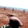 A soldier in camouflage uniform and dark maroon/brown beret is in a prone firing position on a red-dirt shooting range. Multiple human-silhouette paper targets are visible at varying distances across the open field. The scene appears to be a military marksmanship training exercise.