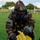 A military personnel wearing full MOPP (Mission Oriented Protective Posture) gear, including woodland camouflage uniform, gas mask, hood, and black gloves, is kneeling on grass while handling a yellow marker. The yellow marker is labeled 'GAS' in red lettering with a red circle, indicating a simulated chemical contamination zone. This appears to be a CBRN (Chemical, Biological, Radiological, Nuclear) training exercise conducted on a military installation.
