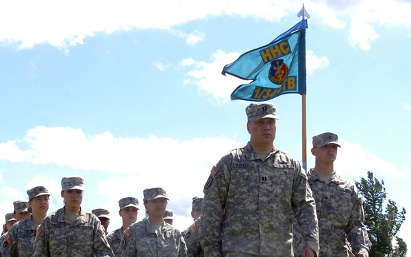A formation of U.S. Army soldiers in ACU (Army Combat Uniform) pattern camouflage are marching across a grass field during what appears to be a formal military ceremony or parade. The lead soldier carries a light blue guidon flag bearing the designation 'HHC 1/34 TB' (Headquarters and Headquarters Company, 1st Battalion, 34th Brigade). Multiple columns of soldiers are visible extending into the background.