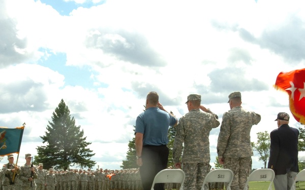 A military ceremony is taking place outdoors on a grassy field, with personnel on a raised platform saluting while formations of soldiers stand at attention in the background. A red flag with three white stars is prominently displayed on the right side of the platform, indicating the presence of a Lieutenant General (3-star general). The event appears to be a formal military review, change of command, or similar official ceremony.