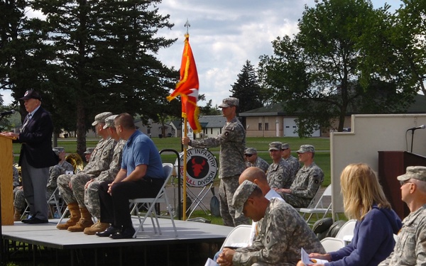 An outdoor military ceremony is taking place on a grassy field at what appears to be a U.S. Army installation. A civilian speaker addresses the audience from a podium on a raised platform, while soldiers in ACU (Army Combat Uniform) and a few civilians are seated in white folding chairs. A unit guidon (orange/red flag) is being held by a soldier, and a banner visible in the background references the 4th Infantry 'Bull' Division.