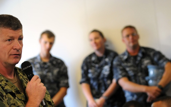A senior military officer in US Navy digital camouflage (NWU Type III) is addressing a group of sailors while holding a microphone. The audience, wearing Navy Working Uniforms (NWU Type I, blue digital pattern), are seated and standing in what appears to be a briefing room or meeting hall. The speaker's name tape reads 'WEST' and he is gesturing with an open hand while speaking.