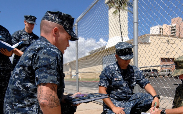 A group of military personnel in U.S. Navy Working Uniform (NWU) digital camouflage are gathered outdoors near a chain-link fence in what appears to be a warm, sunny location. One individual wearing tan/desert camouflage pattern uniform is seated on the ground writing on a notepad or clipboard, while others kneel or stand around him holding documents or folders. The scene suggests a briefing, inspection, or administrative activity taking place outside a secured facility.