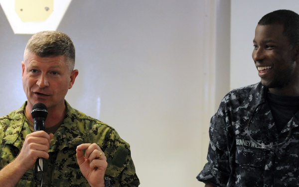 Two U.S. military personnel are standing together in what appears to be an indoor briefing or ceremony setting. The individual on the left, wearing green digital camouflage (NWU Type II/AOR1), is speaking into a microphone. The individual on the right, wearing Navy blue digital camouflage (NWU Type III) with 'U.S. NAVY' visible on the uniform, is smiling broadly.