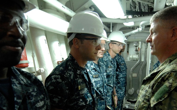 A group of U.S. Navy personnel are gathered in what appears to be a shipboard interior space, likely an engine room or mechanical compartment. Several sailors in Navy Working Uniform (NWU) blue digital camouflage are wearing safety glasses and hard hats, facing a senior officer in green/woodland camouflage uniform. The scene appears to be an inspection, briefing, or walkthrough conducted by the senior officer.