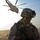 A soldier in full combat gear stands in a desert environment while a large military helicopter hovers in the background. The soldier is wearing MARPAT (Marine pattern) camouflage, a tactical helmet with mounted equipment, and is carrying a rifle. Dust is being kicked up by the helicopter's rotor wash in what appears to be an arid, open terrain.