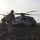 Military personnel in combat gear are disembarking or moving away from a large military helicopter on arid, rocky terrain. The scene appears to be a tactical insertion or extraction operation at either dawn or dusk based on the low-angle backlighting. Multiple soldiers carrying equipment and weapons are visible moving across the landscape.