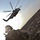 A military service member in full combat gear and desert camouflage is crouching on rocky terrain while a large military helicopter hovers overhead. The image is taken from a low angle looking upward, with strong backlighting creating a dramatic silhouette effect. Dust or haze is visible in the background, suggesting an arid desert environment.