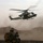 A military helicopter, identifiable as a UH-1Y Venom (Marines variant), hovers low over a dusty, arid desert terrain while rotor wash kicks up significant dust clouds. A ground soldier in full combat gear and a boonie hat crouches in the foreground, facing the aircraft. The scene depicts an active military operation in a desert environment, consistent with operations in Afghanistan or a similar arid region.