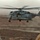 Two military personnel in camouflage uniforms observe a large heavy-lift helicopter landing or hovering in a desert/arid environment. The helicopter appears to be a Sikorsky CH-53E Super Stallion in gray military livery, kicking up dust as it approaches the ground. The scene suggests a tactical insertion, extraction, or resupply operation in a remote, rocky terrain.