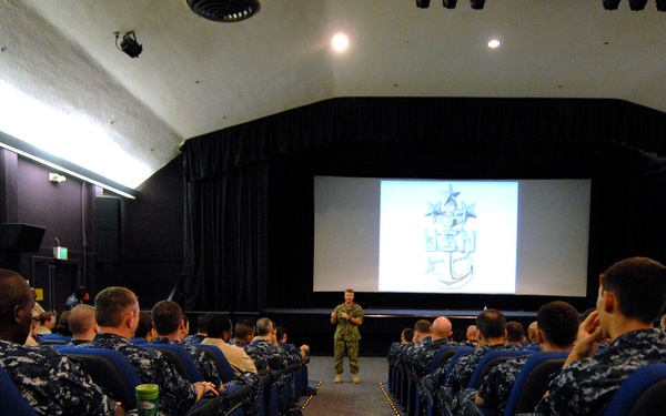 A military briefing or presentation is taking place in a theater-style auditorium. Numerous personnel wearing U.S. Navy digital camouflage (NWU Type I) uniforms are seated in blue theater chairs, facing a speaker standing at the front near a projection screen. The screen displays what appears to be a U.S. Navy Master Chief Petty Officer insignia or related logo.