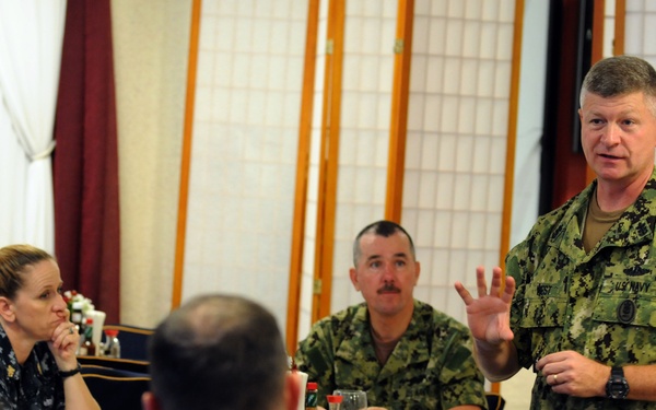 A U.S. Navy officer in digital camouflage (NWU Type III) uniform is standing and addressing a group of military personnel seated around a table in what appears to be a formal dining or briefing room. The seated personnel are wearing various military uniforms including U.S. Navy NWU Type I (blue digital) and what appears to be a Canadian Forces uniform. The setting suggests a formal military meeting, briefing, or working lunch.