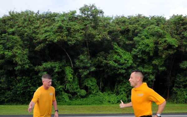 Two men wearing matching yellow shirts and black shorts are on a paved road surrounded by dense green trees. One man is actively running while the other stands and watches. Both appear to be participating in a physical fitness activity, likely a military or organized PT (physical training) session based on their matching uniforms.