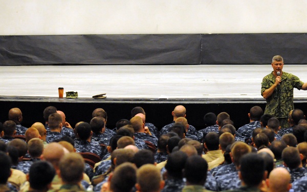A military briefing or all-hands meeting is taking place in what appears to be an auditorium or theater. A single speaker in camouflage uniform stands on a stage holding a microphone and gesturing, addressing a large audience of seated military personnel wearing various uniforms. The crowd appears to number in the hundreds.