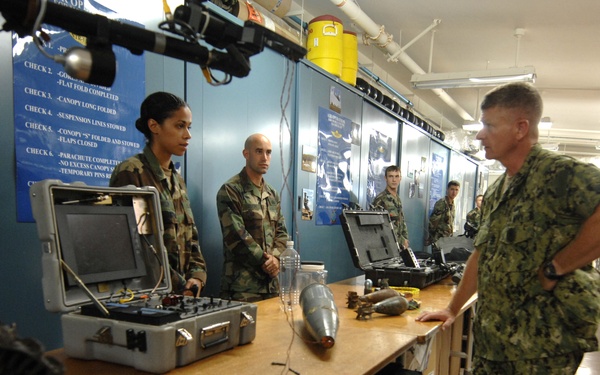 Military personnel in camouflage uniforms are gathered in what appears to be a training or equipment room, examining items on a workbench. A senior Navy officer in digital camouflage (NWU) is being briefed or shown equipment by junior personnel. The room contains various military equipment including a portable case with electronics, projectile-like objects, and what appears to be a robotic device on the left.