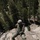 1/1 Marines Begin Exercise Mountain Warrior at Bridgeport A military service member in MARPAT (Marine pattern) camouflage uniform is rappelling down a rocky cliff face in a mountainous, forested environment. The individual is equipped with a helmet, gloves, harness, and rappelling rope, suggesting a military mountain warfare or combat training exercise. At least two other personnel are visible in the background on the rock face.