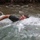 1/1 Marines Begin Exercise Mountain Warrior at Bridgeport A young male is actively swimming through turbulent water, likely a river or stream, with significant water splashing around him. He is wearing a dark shirt and appears to be exerting considerable effort, with his face turned sideways to breathe. The background shows murky greenish water and what appears to be vegetation or debris along the bank.