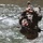 1/1 Marines Begin Exercise Mountain Warrior at Bridgeport A military service member in full combat gear is traversing a rope line over a fast-moving river, suspended upside down while gripping the rope with both hands. The individual is wearing MARPAT (Marine pattern) camouflage uniform, suggesting affiliation with the United States Marine Corps. This appears to be a military training exercise involving a water obstacle crossing technique.