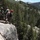 1/1 Marines Begin Exercise Mountain Warrior at Bridgeport Military personnel in camouflage uniforms are conducting rappelling/rock climbing training on a granite cliff face in a mountainous, forested environment. One individual wearing a red safety helmet and harness stands at the cliff edge overseeing the exercise, while another soldier is positioned lower on the rock face connected to a rope. The backdrop features dense conifer forests, rocky terrain, and snow-capped mountain peaks consistent with a high-altitude training environment.