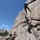 1/1 Marines Begin Exercise Mountain Warrior at Bridgeport Military personnel in camouflage uniforms are conducting a rock climbing or rappelling training exercise on a large granite rock face. At least one climber is ascending the rock wall using ropes while two or three personnel at the base manage the belay lines. The setting appears to be a mountainous environment with snow-capped peaks visible in the background.