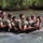 1/1 Marines Begin Exercise Mountain Warrior at Bridgeport A group of approximately 8-9 individuals wearing olive green military-style t-shirts are wading through a river or stream in a tight formation. They appear to be carrying a log collectively, suggesting a team-building or military training exercise. The setting is an outdoor waterway with green vegetation visible along the bank in the background.
