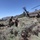 Military personnel in combat uniforms are carrying a casualty on a litter (stretcher) toward a CH-47 Chinook helicopter in a mountainous, high-desert terrain. The scene depicts a medical evacuation (MEDEVAC) or CASEVAC operation, likely during a training exercise. The helicopter's rotors are spinning and it appears to be on or near the ground awaiting the casualty.